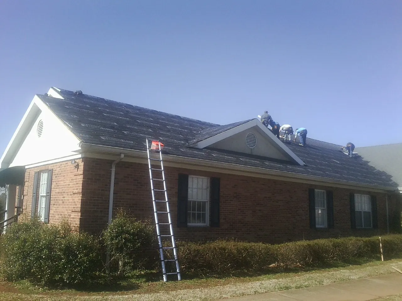 Crew tearing shingles off a brick ranch roof with a ladder set — Emergency Roof Repair work by our crew in Rowena