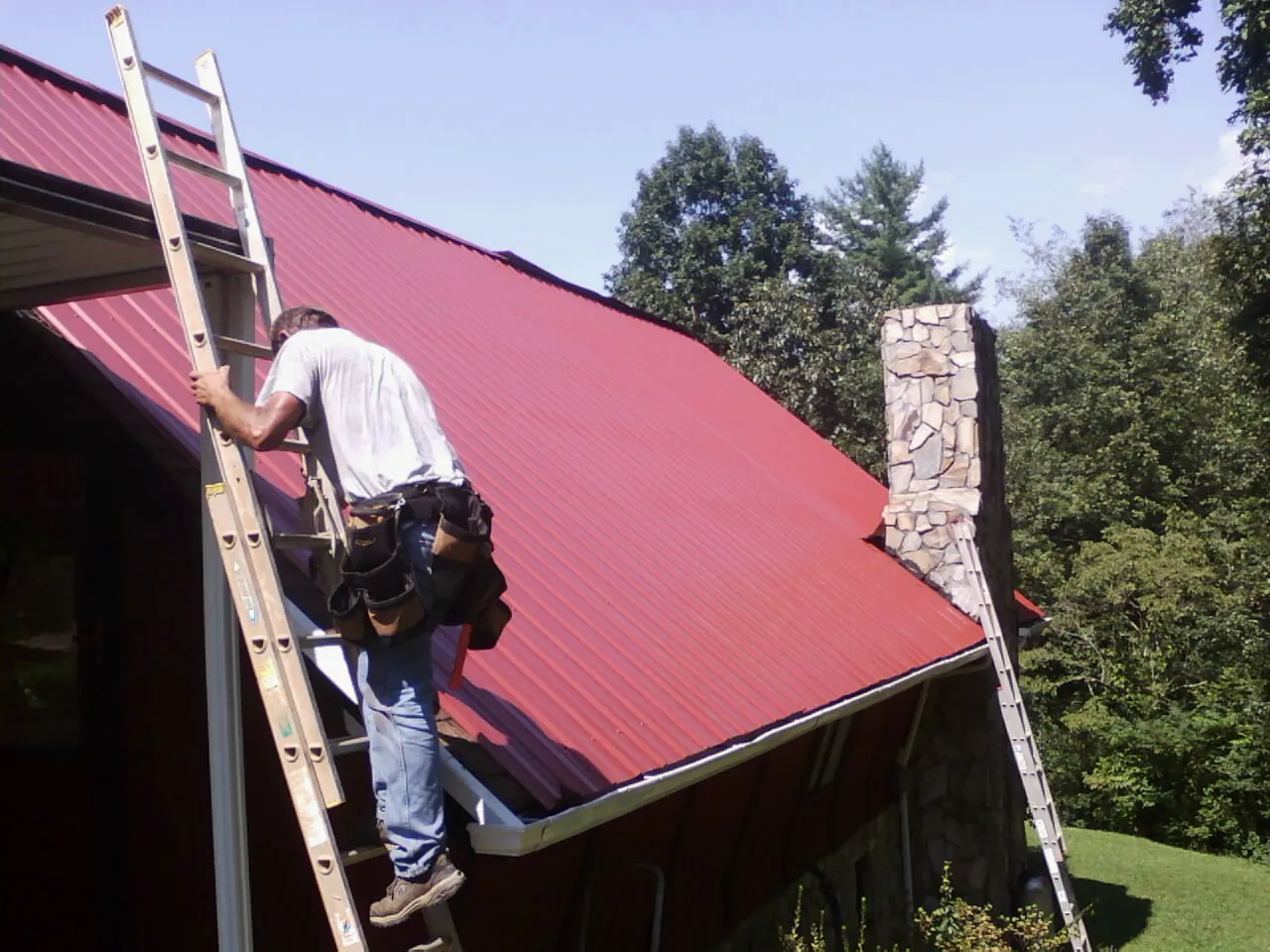 Roofer on a ladder beside a red metal roof and stone chimney — Soffit & Fascia Repair by Roofing Brothers in Rowena